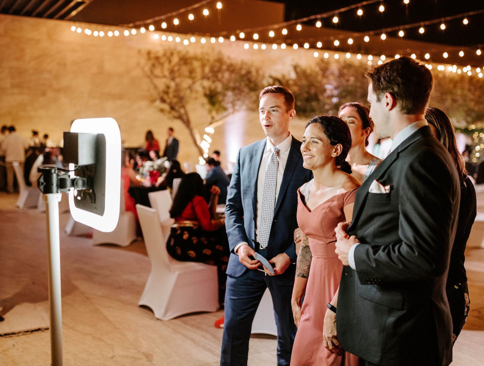 Des invités en tenue de soirée rient devant le vidéobooth, sous des guirlandes lumineuses